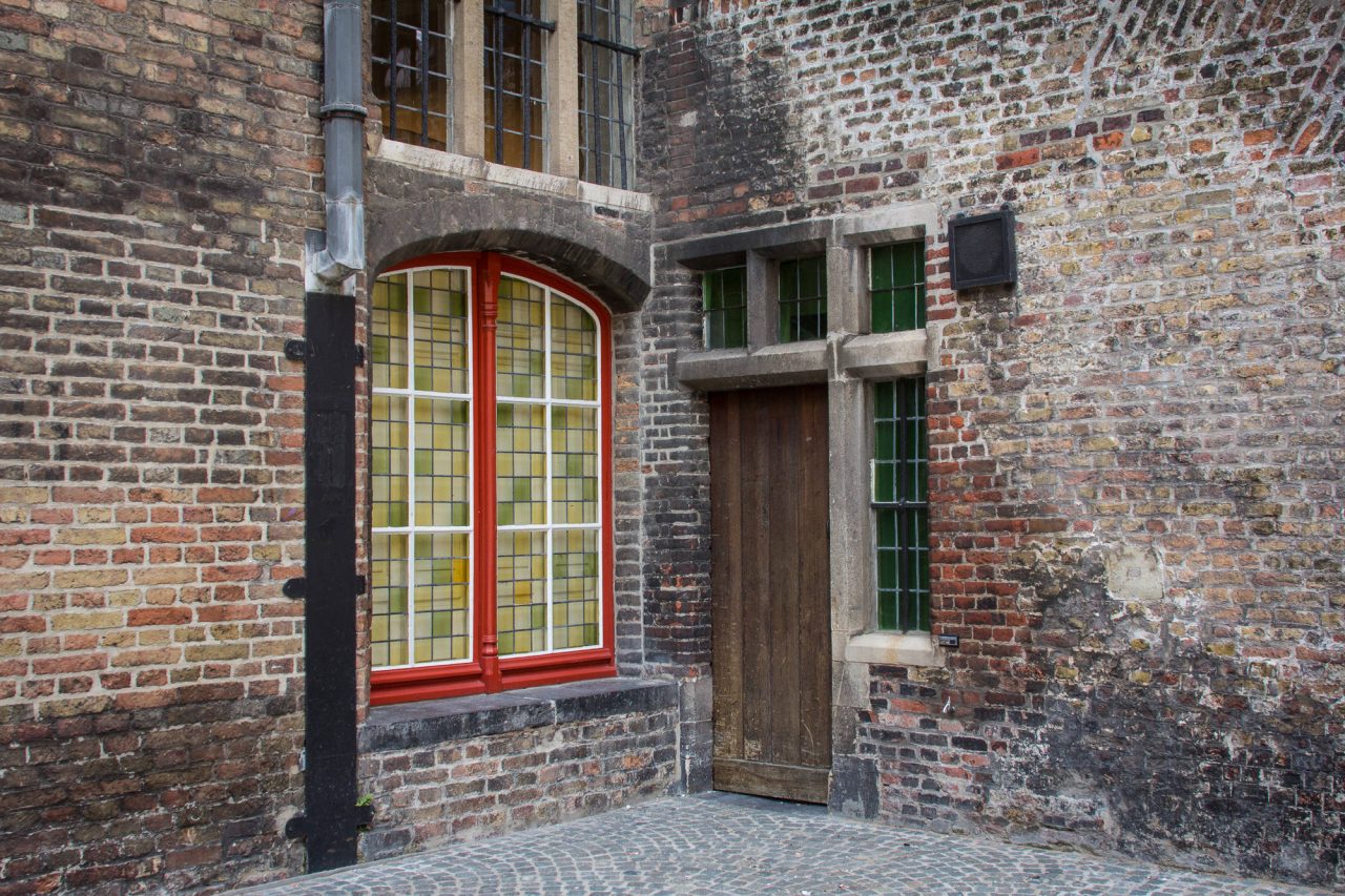 A weathered brick façade with a bright red window frame and wooden door